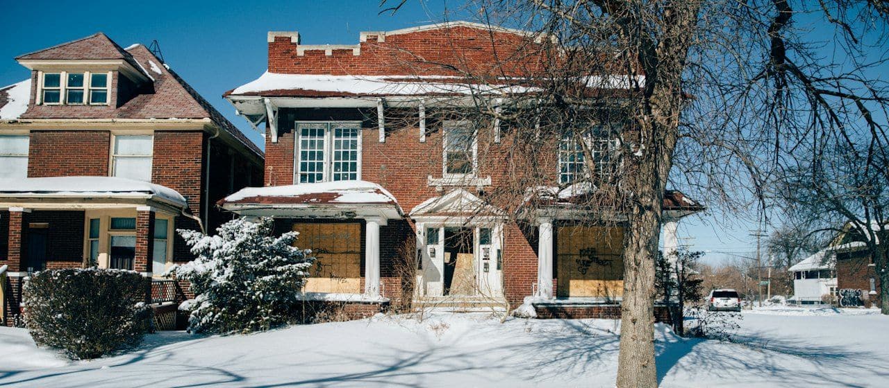 Street-level photo of a boarded-up brick house in the snow